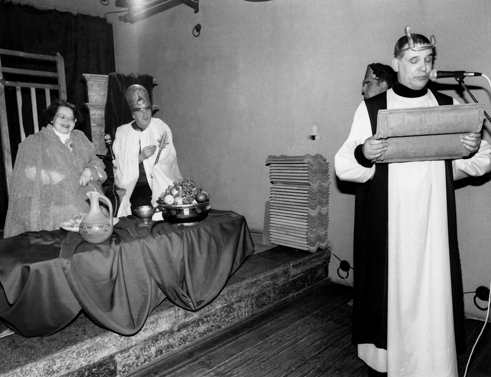 Augusts Sukuts (no labās) lasa svinīgo apsveikuma runu no māla plāksnītēm, svinot Valentīnas Freimanes un Jura Stiprā 75 gadu jubilejas (kopā 150) restorānā "Andalūzijas suns" 1997. gadā