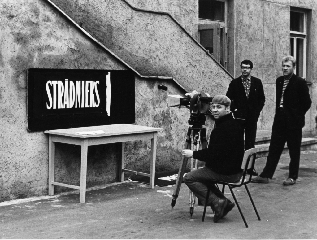 Režisors un operators Uldis Brauns Rīgas kinostudijas pagalmā filmē titrus savas triloģijas trešajai filmai Strādnieks (1963), fonā stāv triloģijas scenārists Armīns Lejiņš. Foto no Rīgas Kino muzeja krājuma